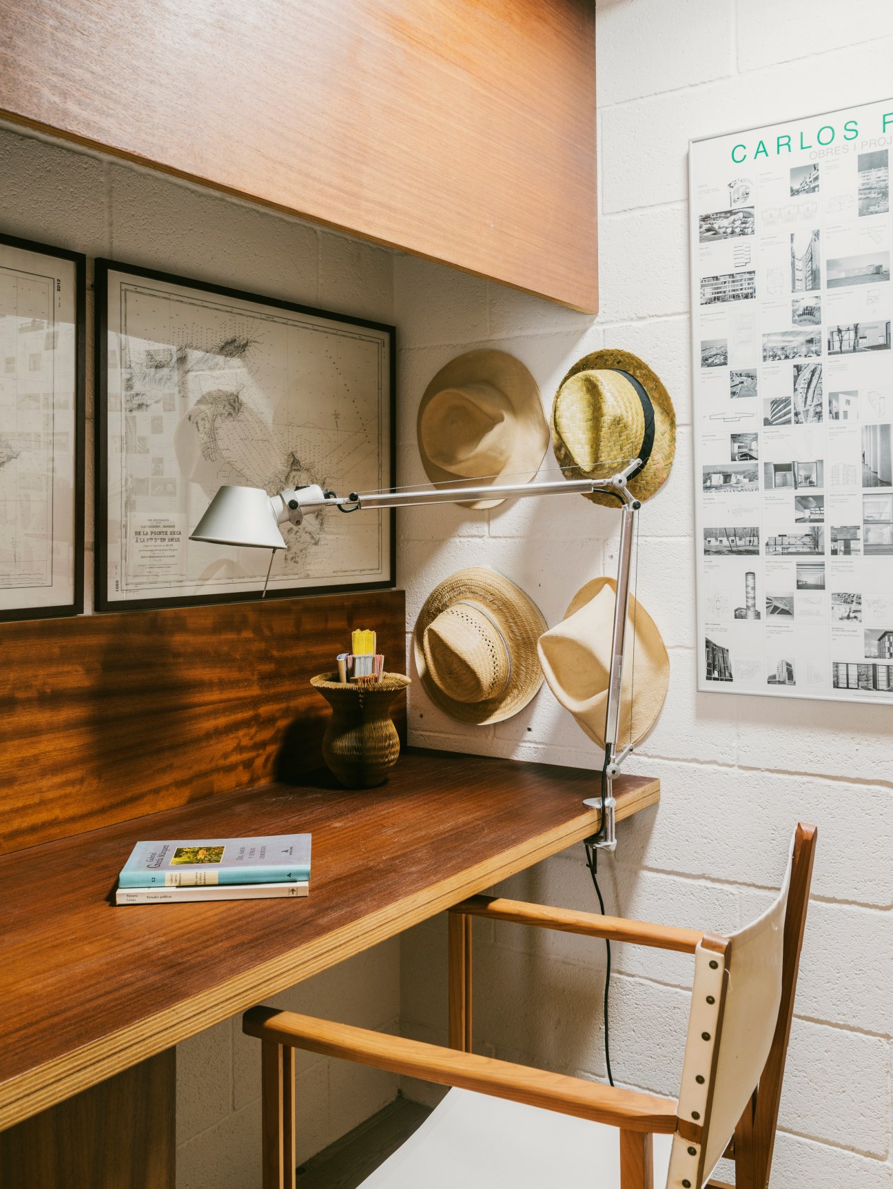 Architect Carlos Ferrater's minimalist workspace with wooden desk, framed architectural drawings, articulated desk lamp and straw hats on wall.
