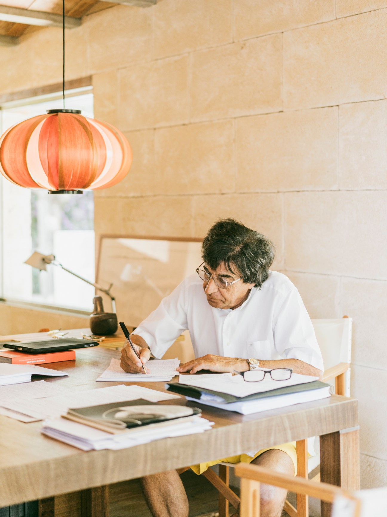 Architect Carlos Ferrater works at a wooden desk in a light-filled studio, with an orange Coderch pendant lamp above.