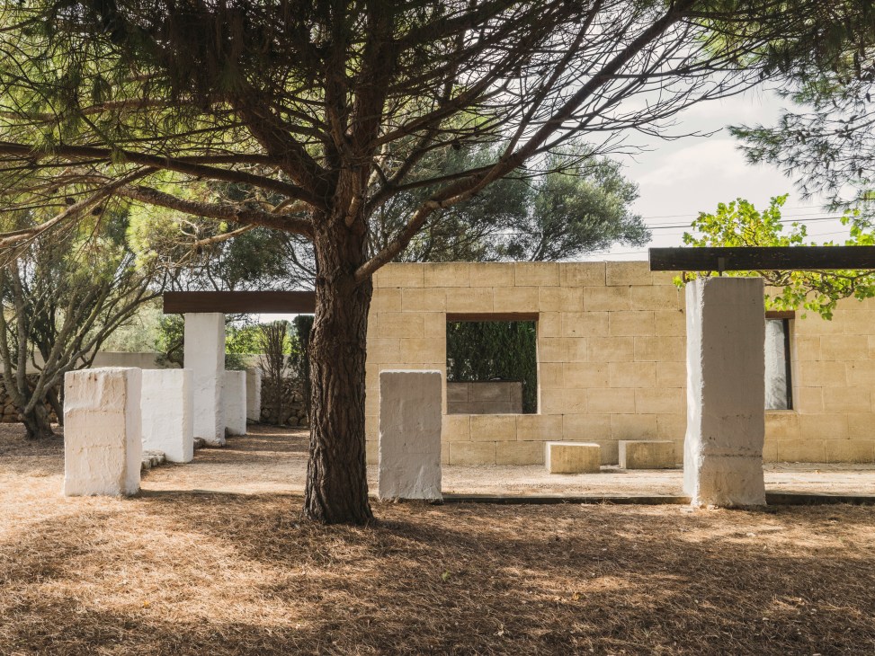 A modern stone pavilion with white pillars under a large tree in Menorca, referencing ancient Talaiots architecture.