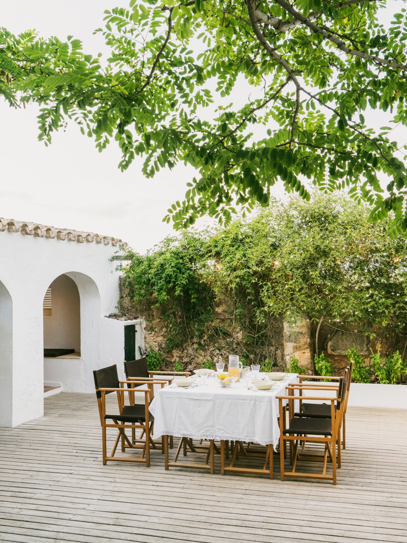 A wooden dining table set for lunch on a deck beside a white-walled stone building with ivy-covered walls and overhanging tree branches.