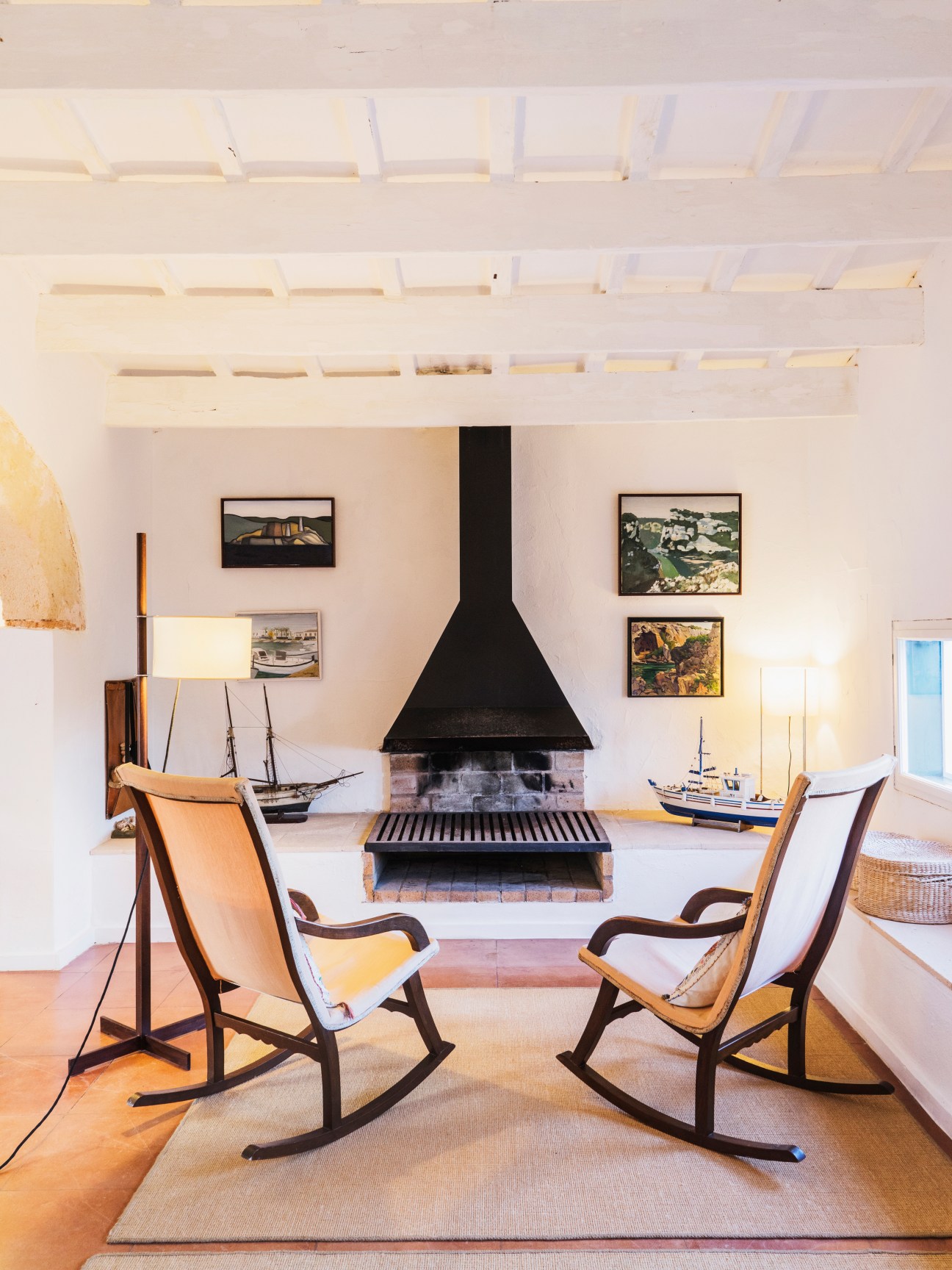 Interior room with black suspended fireplace, wooden rocking chairs, exposed beam ceiling, and framed nautical artwork on white walls.