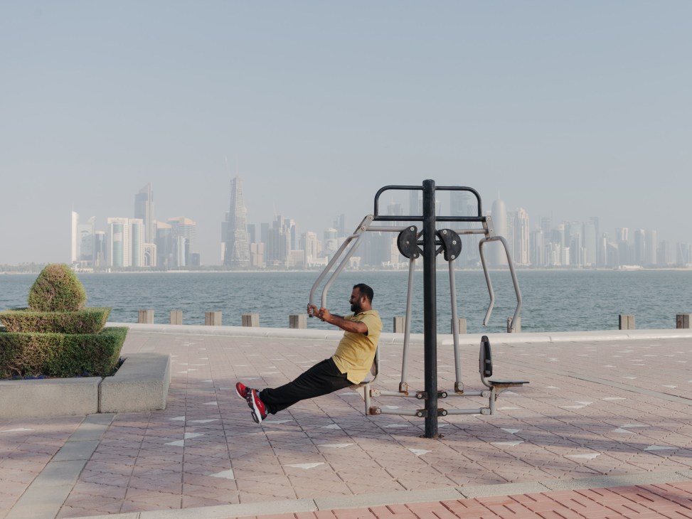 Man exercising on outdoor pull-up equipment in a waterfront park with city skyline in background.