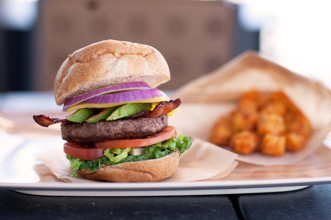 A gourmet burger with whole wheat bun, lettuce, tomato, avocado, bacon, and beef patty on a white plate with roasted vegetables blurred in background.