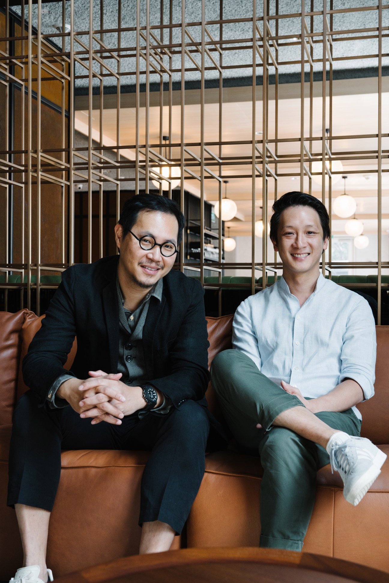 Asylum founder Chris Lee and Wee Teng Wen seated on brown leather sofa against geometric brass screen partition.