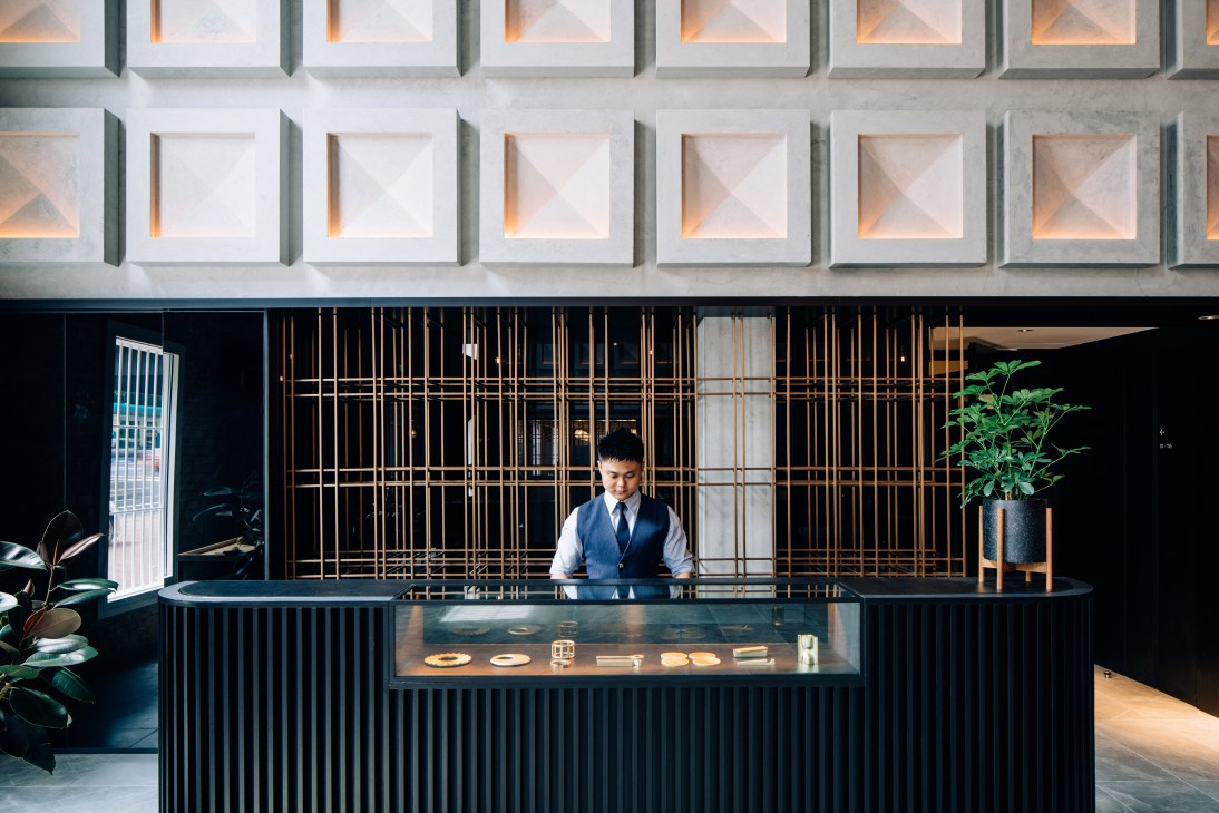 A receptionist stands behind a modern black desk with brass grid screening and geometric ceiling lights above.