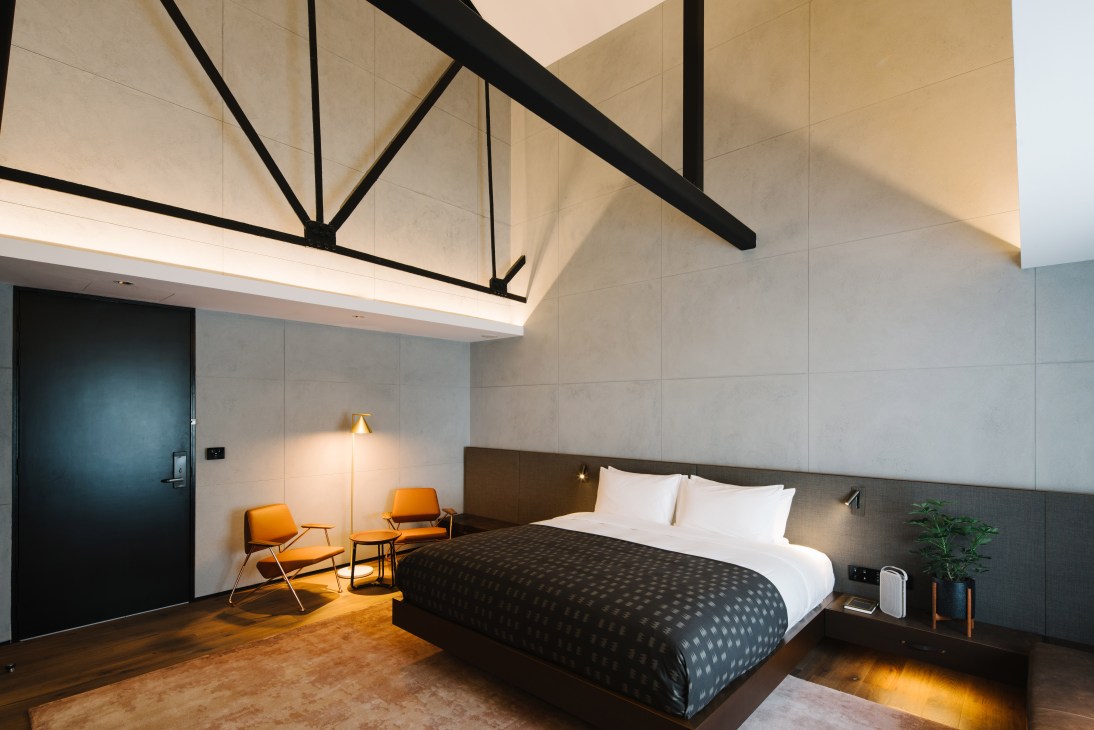 A modern hotel bedroom with exposed black beam ceiling, queen bed with white pillows, orange chairs, and minimalist design elements at The Warehouse Hotel Singapore.