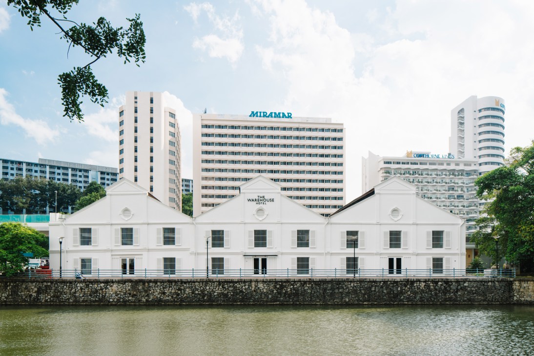 The Warehouse Hotel's white facade with pitched roofs on the Singapore River, surrounded by modern high-rise buildings.