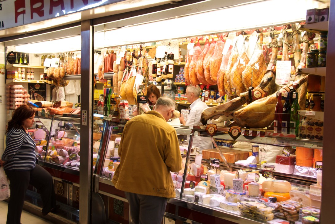 Customers browse a Spanish butcher shop window displaying cured meats, fresh cuts, and market products.
