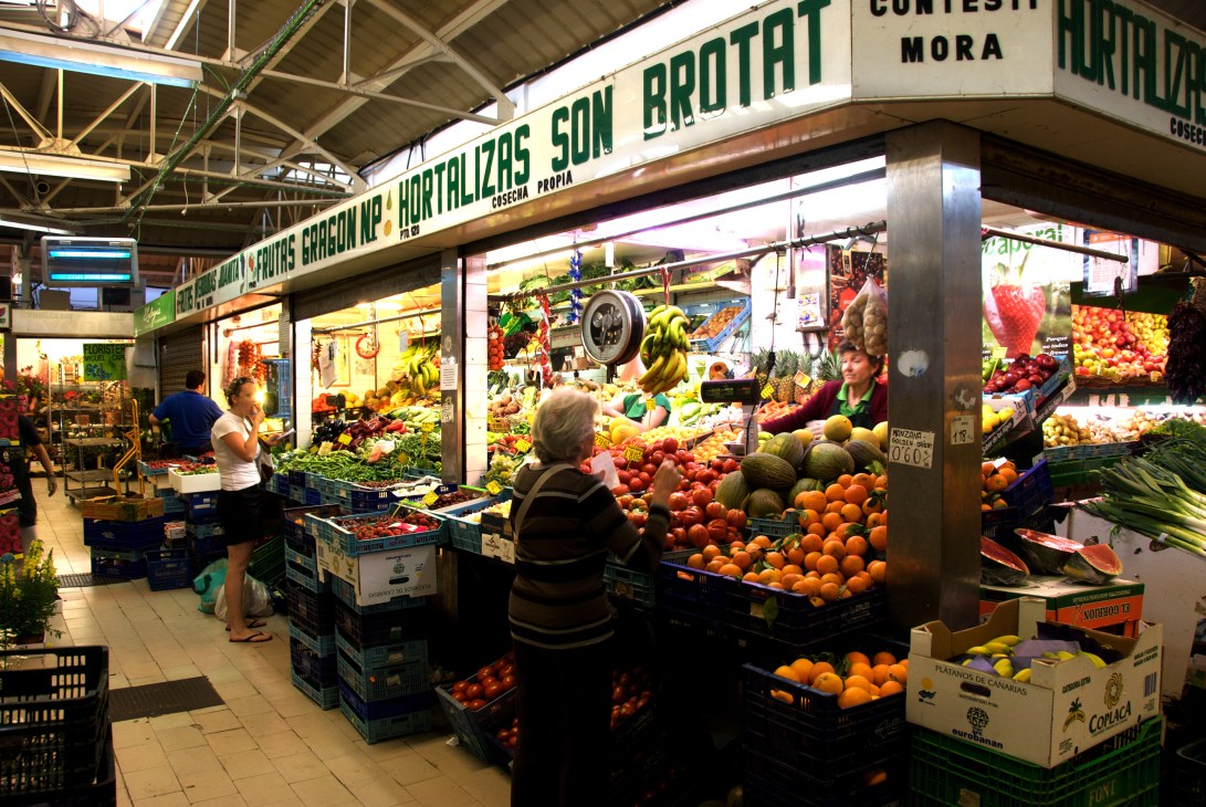 Interior of covered market in Palma de Mallorca with produce vendor stalls displaying fresh fruits and vegetables.