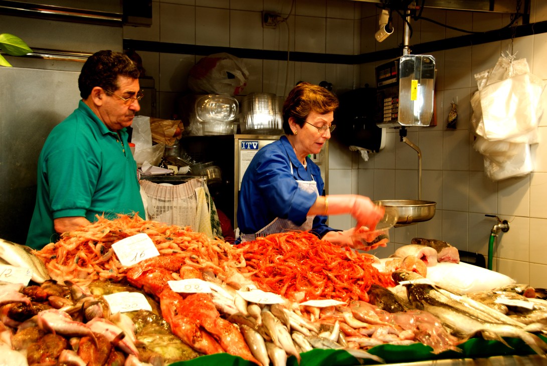 Two vendors at a fish and seafood counter in Palma de Mallorca's covered market display various fresh catches including red shrimp and whole fish.