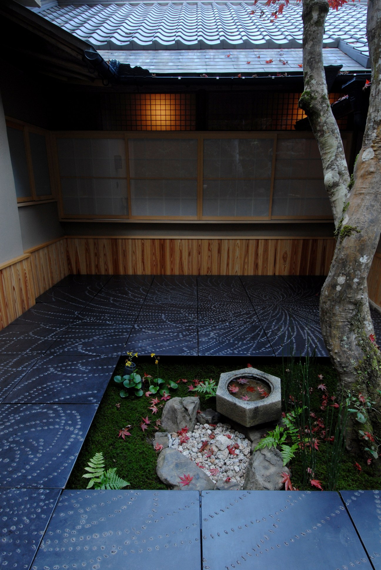 Traditional Japanese ryokan courtyard with moss garden, stone water basin, and wooden architecture with blue-tiled roof.