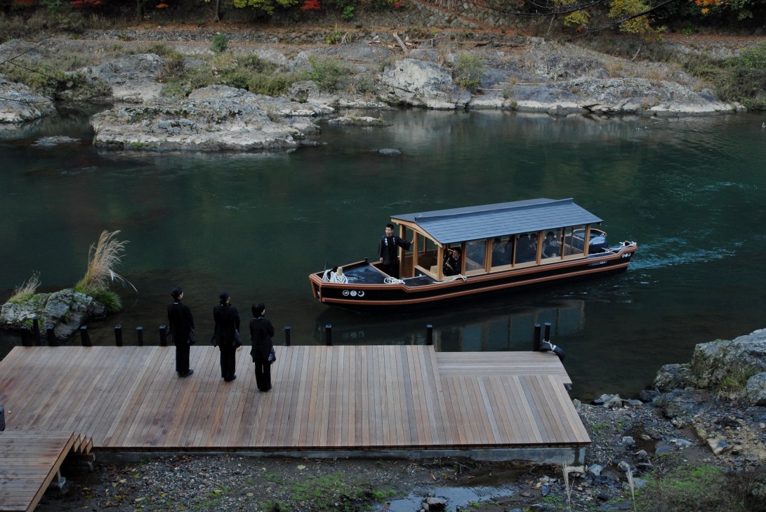 Staff in dark uniforms stand on a wooden dock beside a traditional Japanese wooden boat on a river.