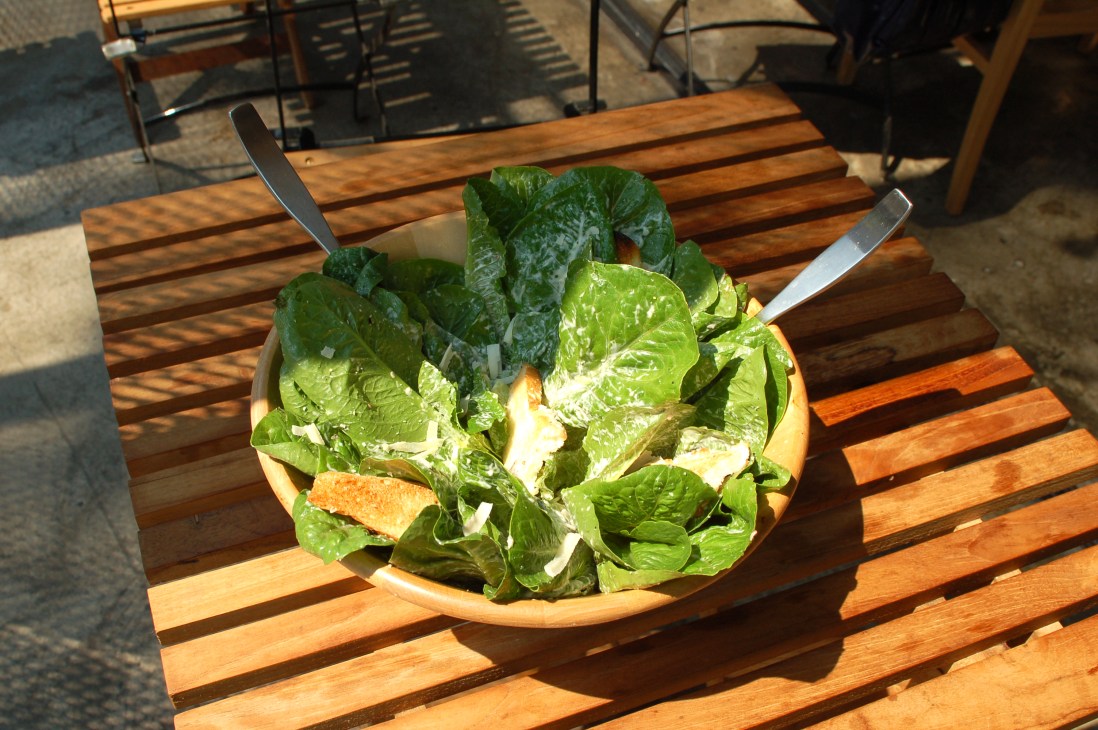 Fresh green salad with mixed lettuce leaves and white cheese on a wooden table in sunlight.