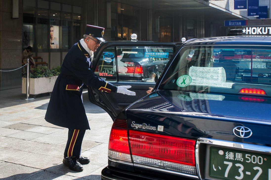 Uniformed taxi attendant in navy and gold uniform opening car door outside Isetan department store in Shinjuku.