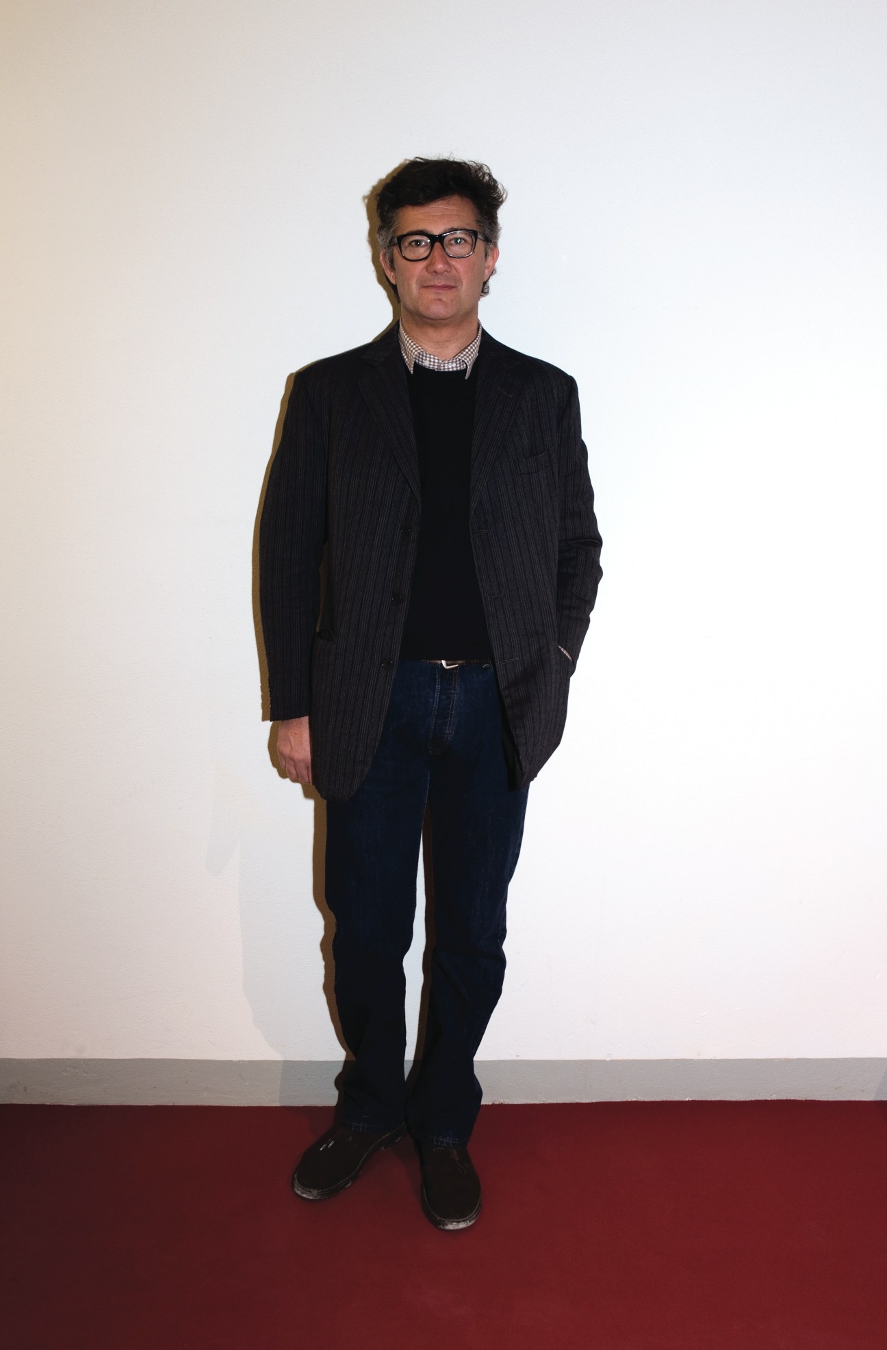 Massimo Piombo, designer, wearing a pinstriped blazer, black sweater, checked shirt, dark jeans and black shoes.
