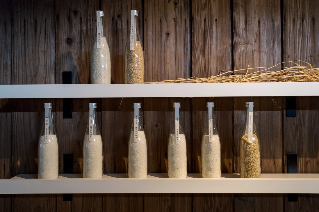 Glass bottles filled with rice grains displayed on wooden shelves at Imayotsukasa saké brewery.