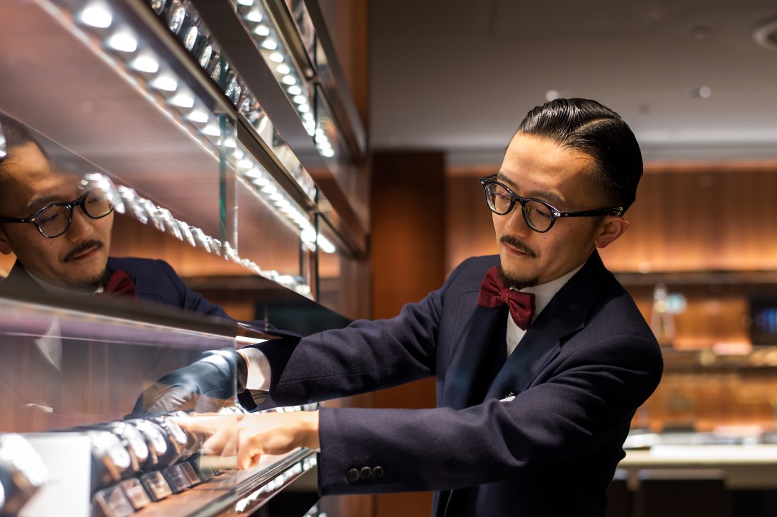Isetan staff member in formal attire and white gloves examining men's watches in an illuminated display case.