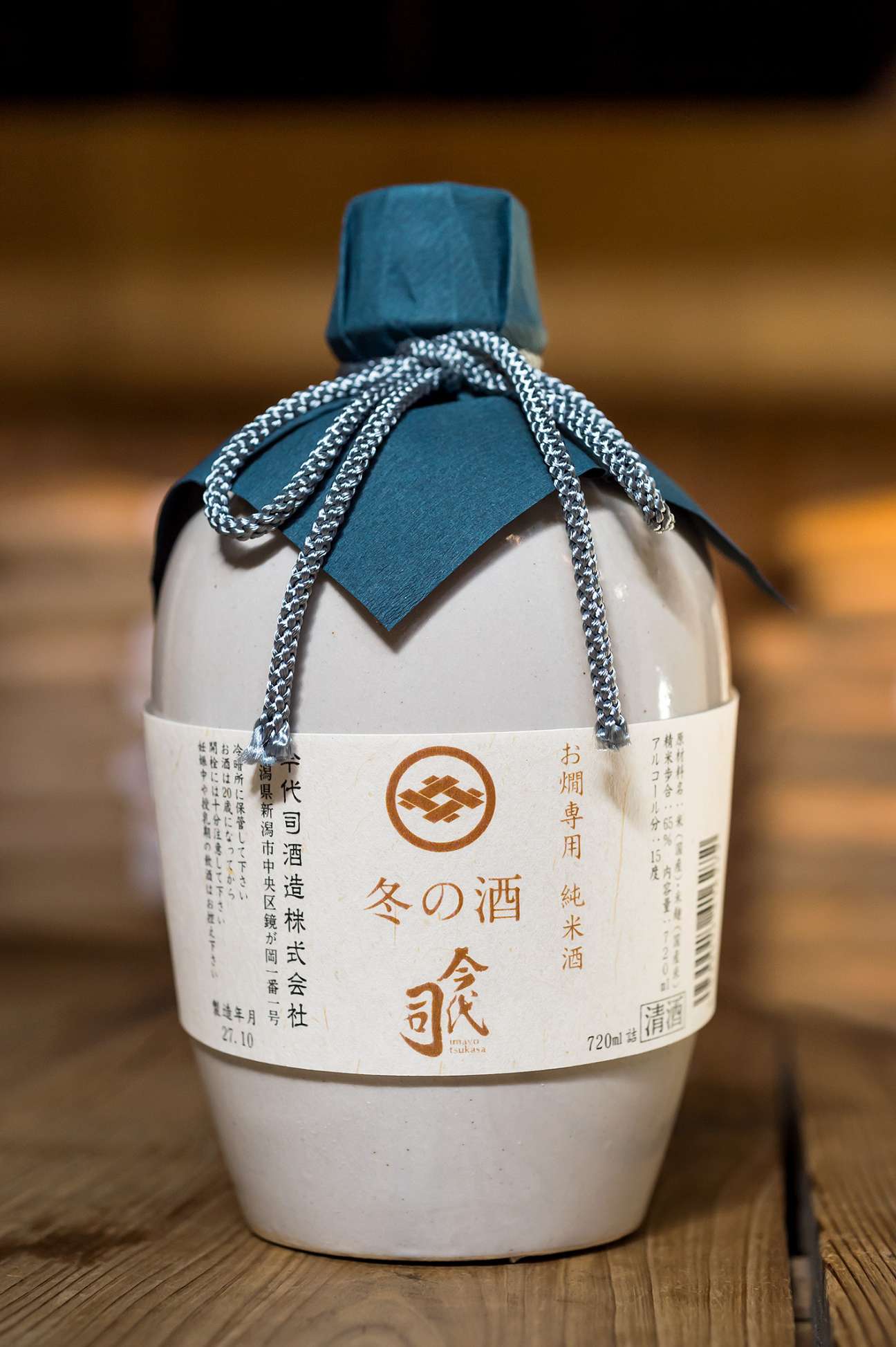 Bottle of Imayotsukasa winter saké with blue cloth stopper and silver cord on wooden surface.