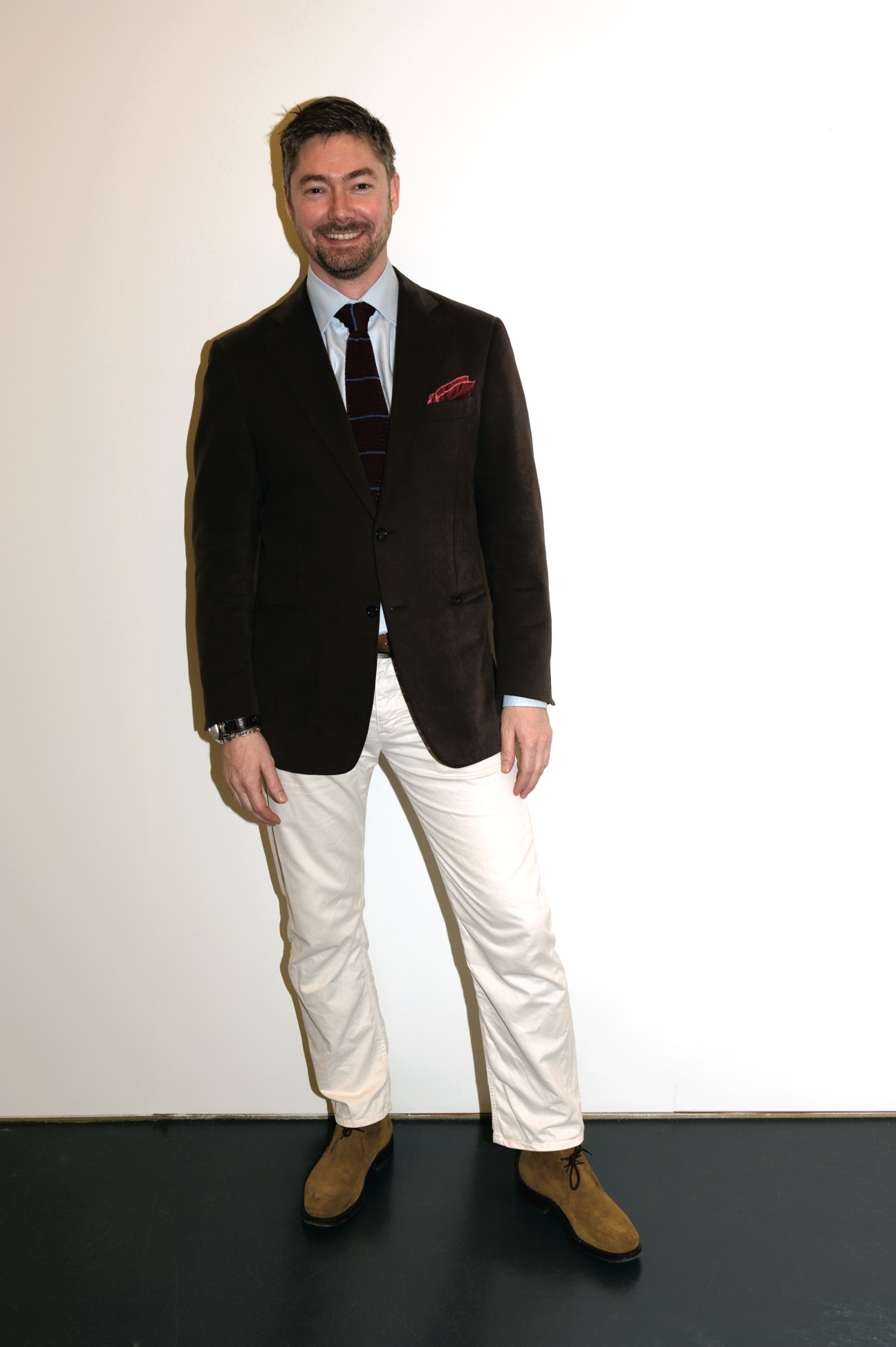 Man wearing black Canali blazer, white trousers, Church's shoes and tie against white background.
