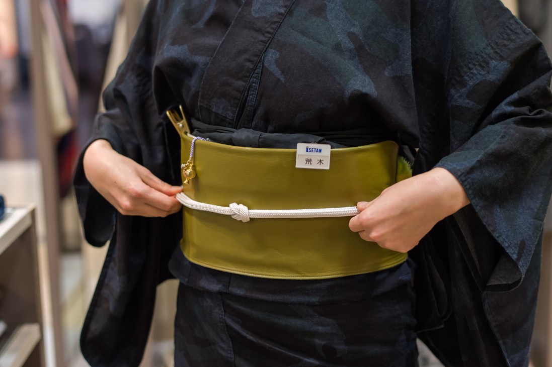 Person wearing black denim jacket holding a mustard-yellow belt with white rope detail and ISETAN label.