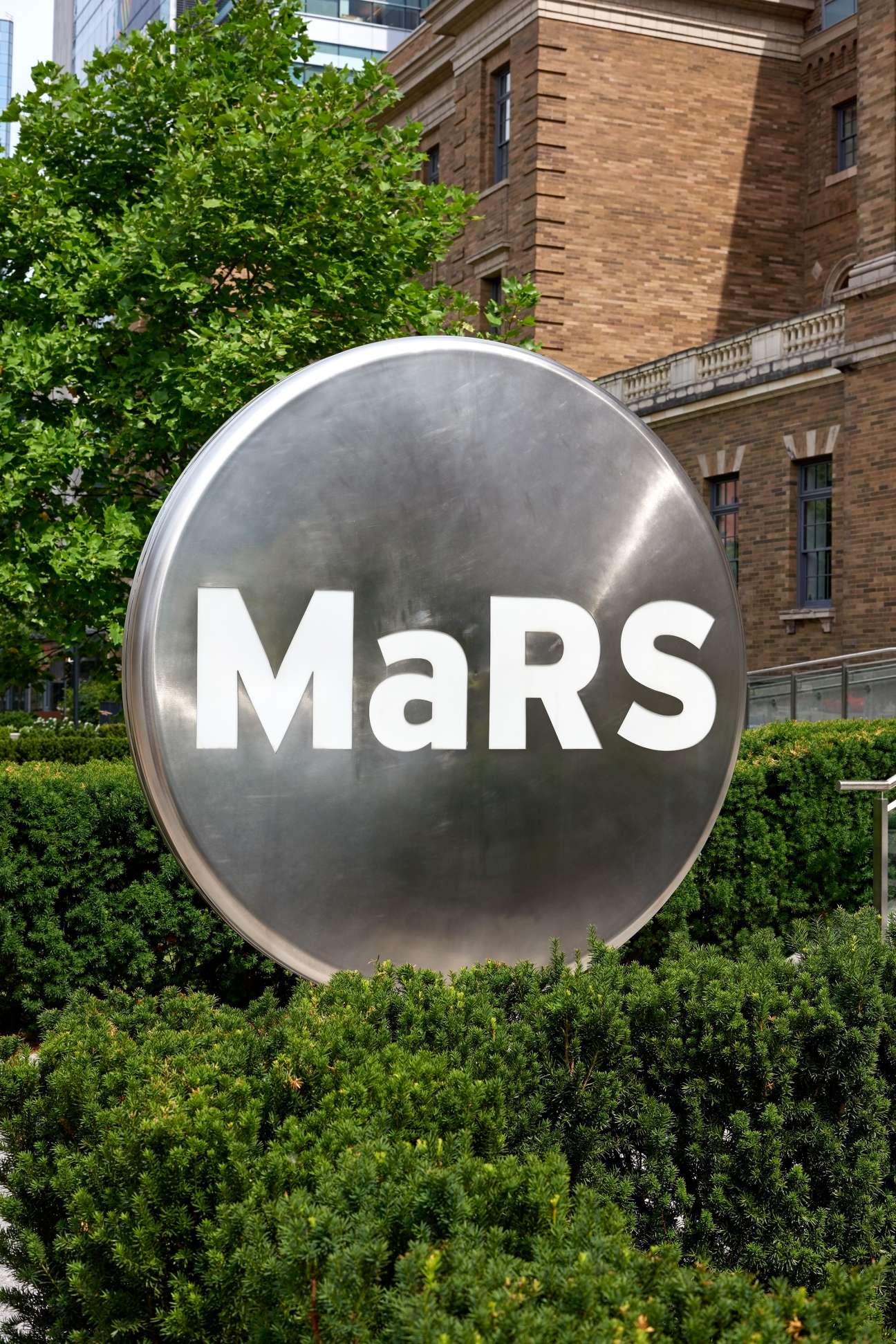 Large metallic spherical sign reading 'MaRS' outside a brick building surrounded by greenery.