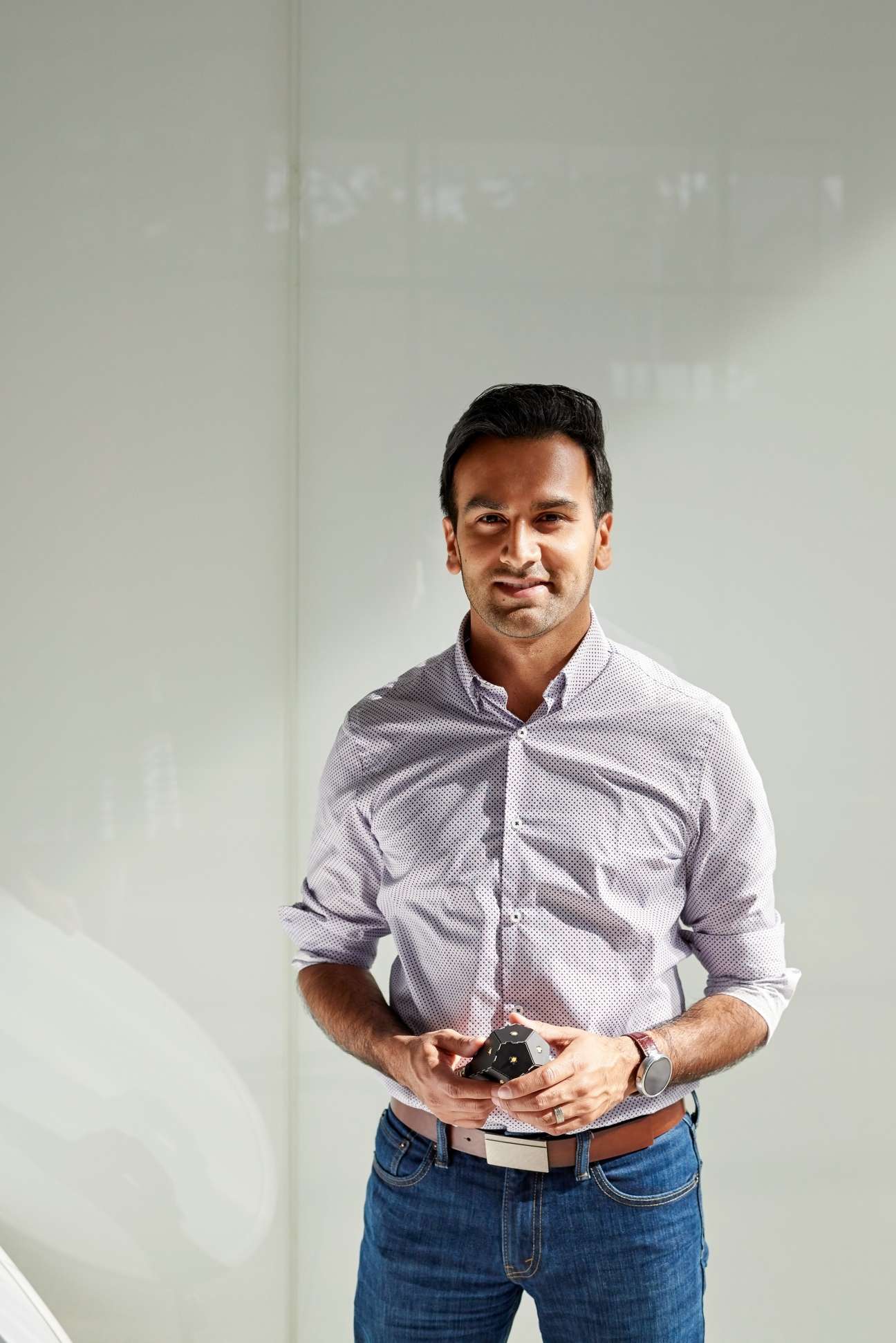 Man in light purple patterned shirt holding a black device against white background