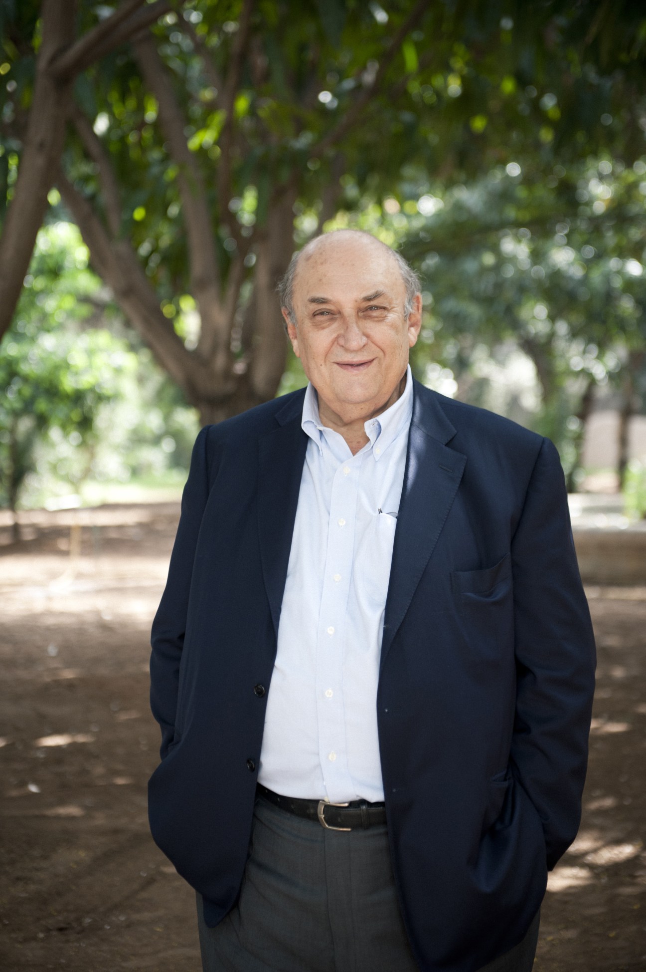 David Corm stands in a tree-lined garden wearing a dark blazer and light blue shirt, smiling at the camera.