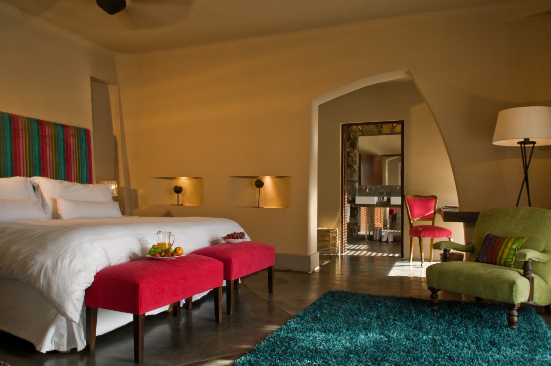 Hotel bedroom with colorful furnishings including red benches, green chair, and striped headboard.
