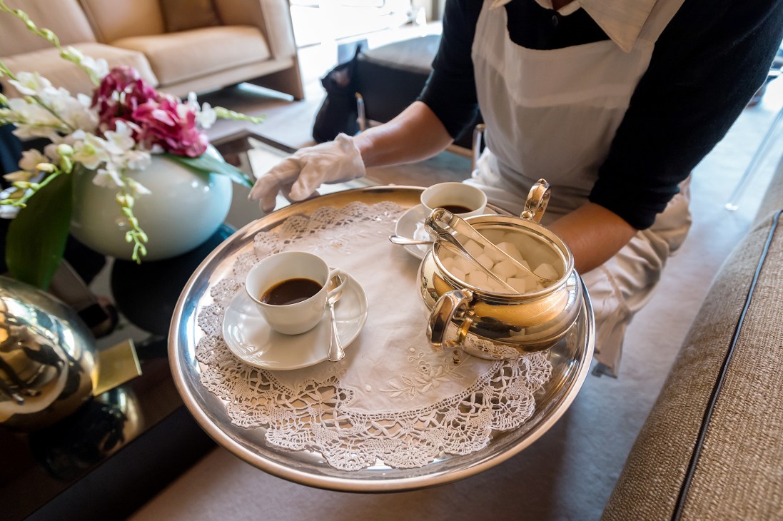 A server in beige apron pours coffee from a gold teapot onto a silver tray with lace doily, white cup and saucer.