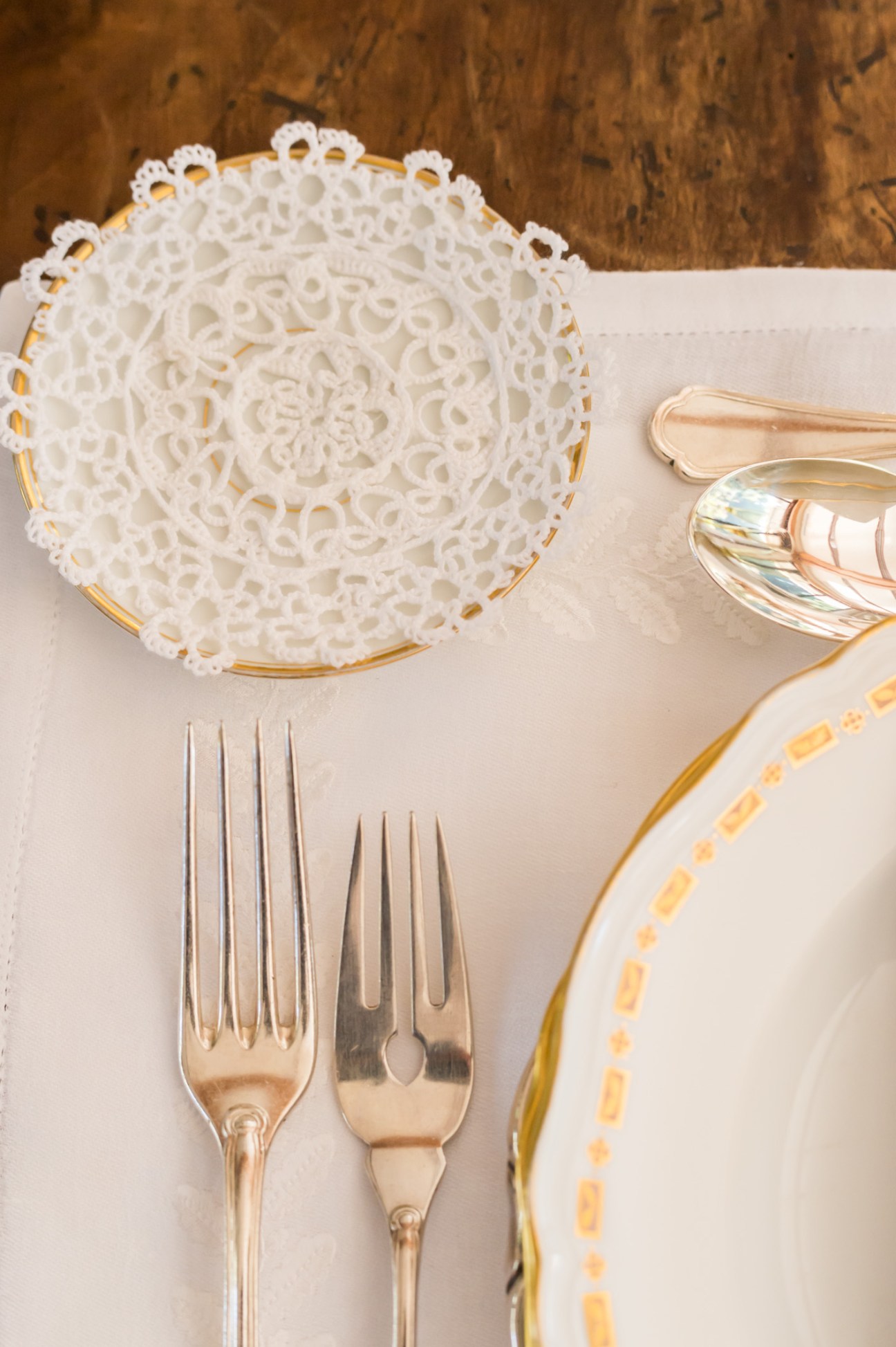 Antique silverware and decorative white lace doily on cream Frette table linen with gold-trimmed plate.