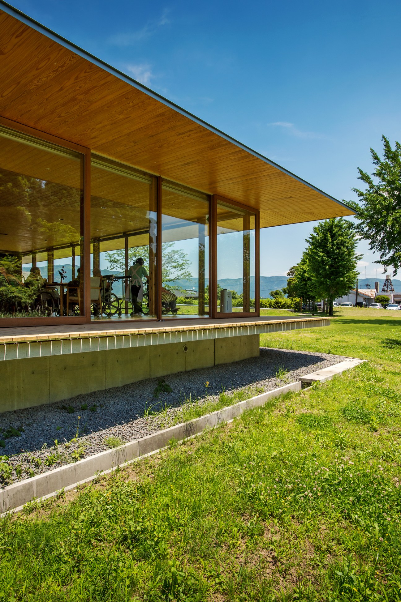 Modern wooden nursing home with floor-to-ceiling windows and covered terrace overlooking green grounds and distant mountains.