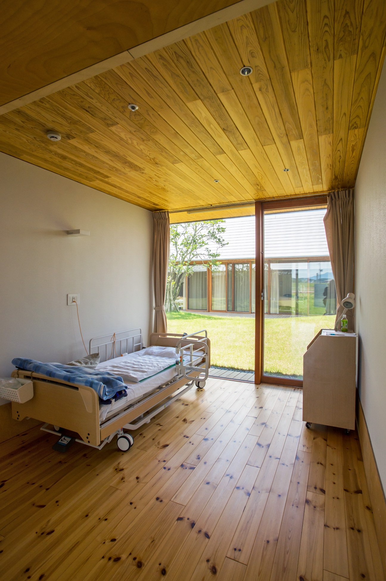 A bright bedroom at Ebino Ryofuen nursing home with wooden ceiling and floor, overlooking an inner courtyard garden through large glass doors.