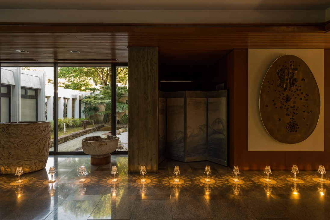 Modern interior hallway with large oval artwork on yellow wall, small golden table lamps along floor, and garden view through windows.