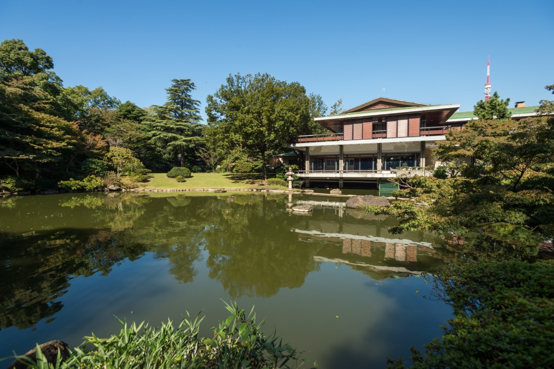 Modern residence with red brick upper level beside a pond, surrounded by manicured gardens and trees.