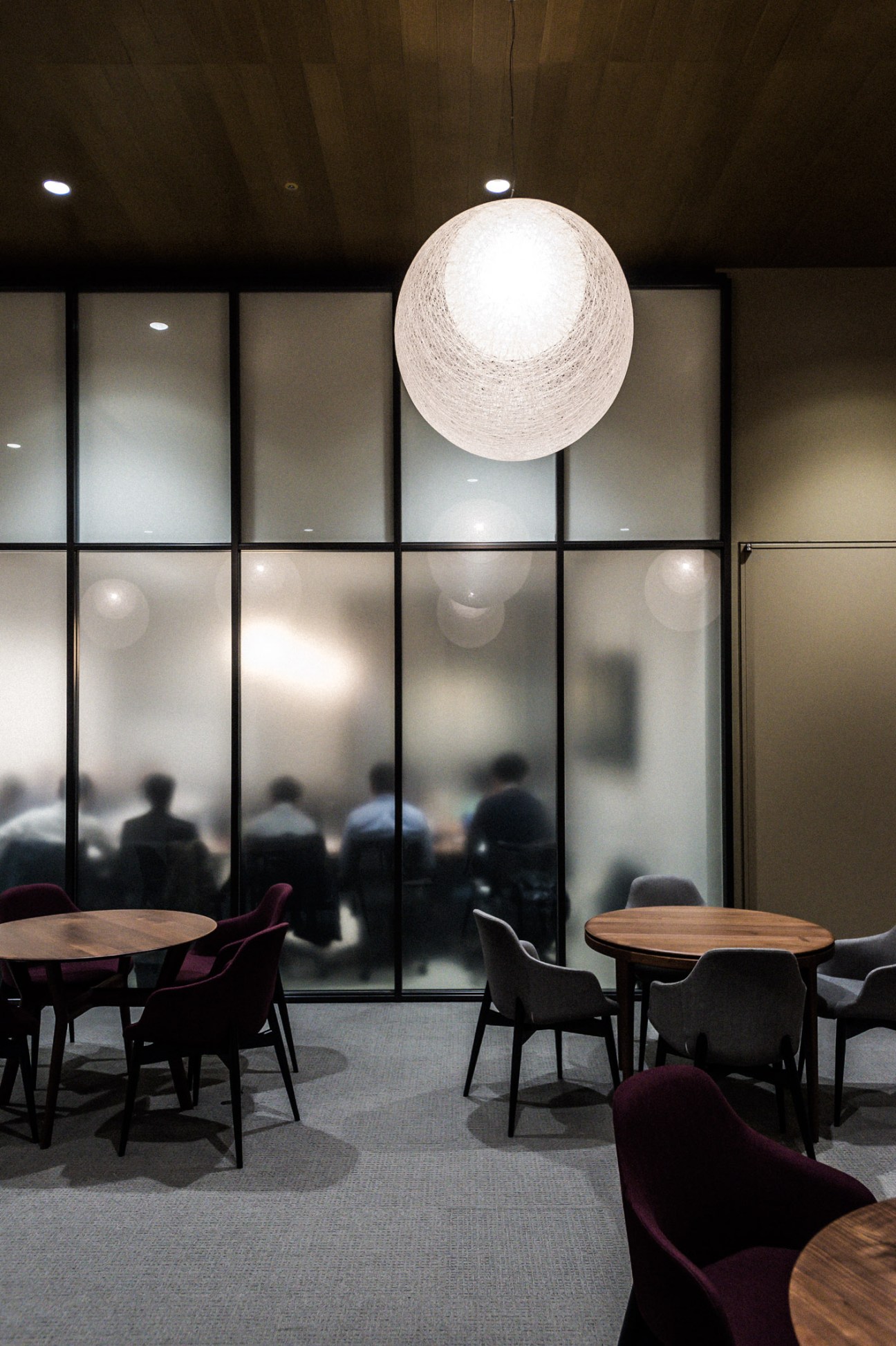 Modern office lounge area with round pendant lamp, wooden tables, and black chairs overlooking Tokyo through floor-to-ceiling windows.