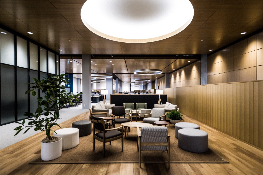 Modern lounge area with wooden flooring, contemporary furniture, potted plants, and a large circular ceiling light in Uniqlo City headquarters.