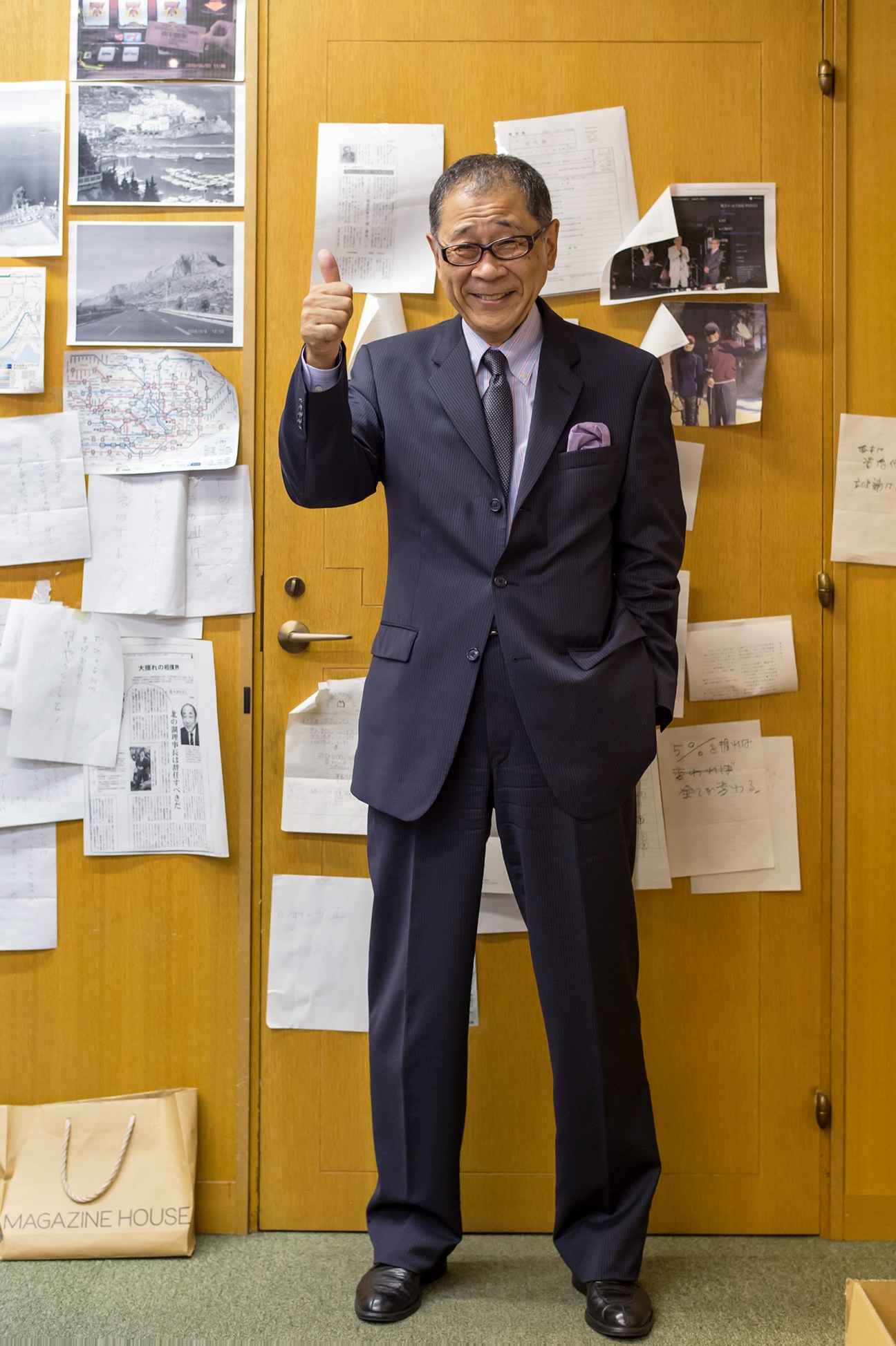 Tsutomu Ishizaki, president of Magazine House, giving a thumbs-up in his office with photographs and documents pinned to yellow walls behind him.