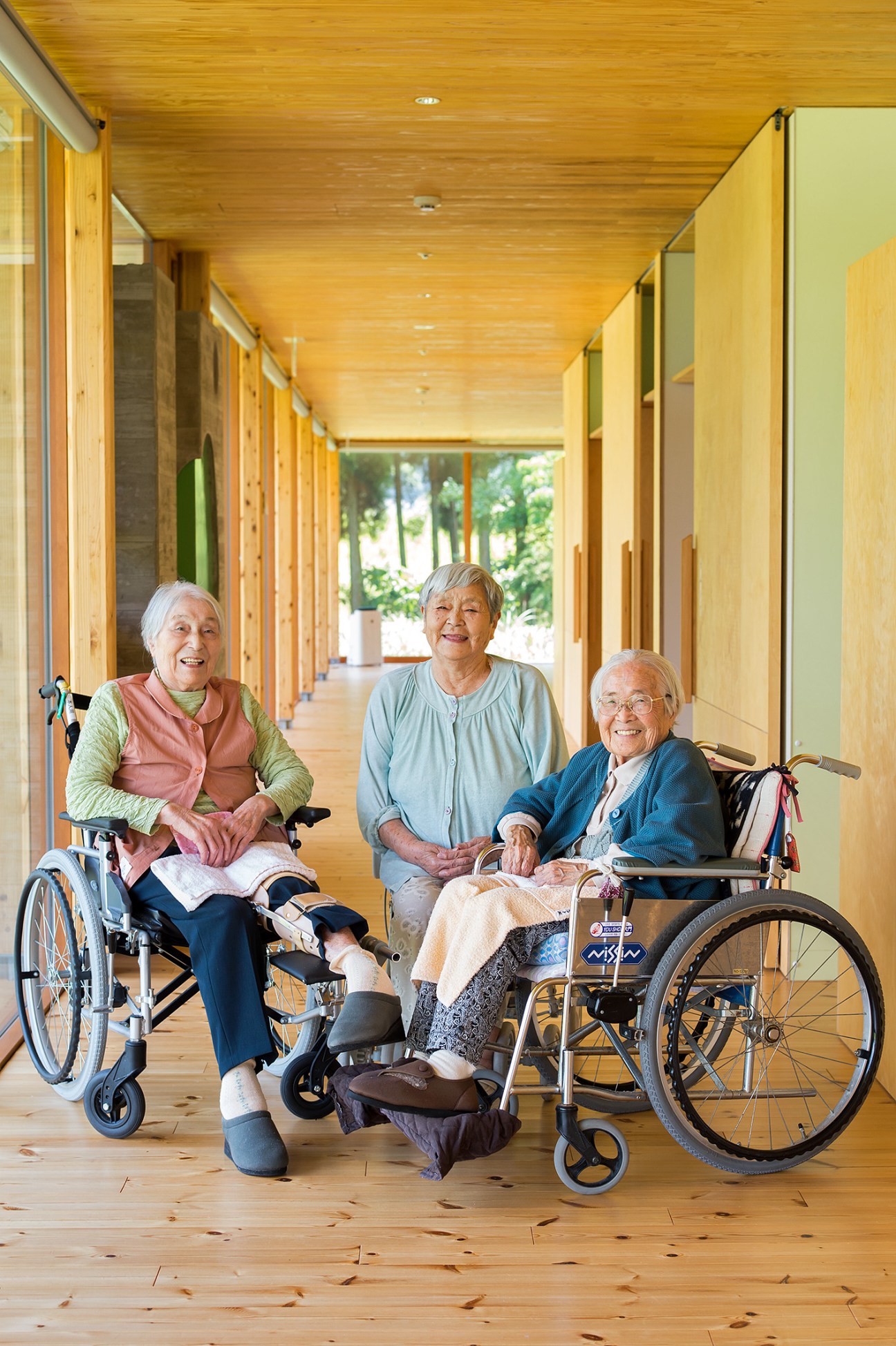 Three elderly women in a wooden corridor at Ebino Ryofuen nursing home, two in wheelchairs smiling at camera.