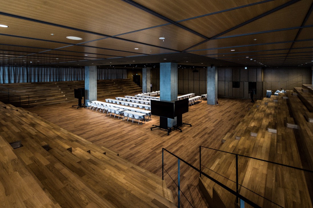 A large modern conference hall with wooden flooring, white chairs arranged in rows, and a wood-paneled ceiling with integrated lighting.
