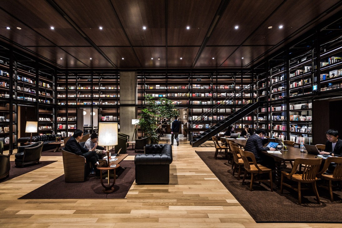 Modern library interior with floor-to-ceiling black shelving units filled with books, wooden flooring, and staff members working at tables.