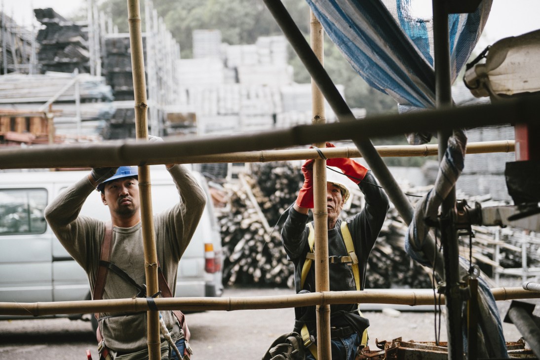 Two bamboo scaffolders, Tang and Yu, tying knots while working on a bamboo scaffold structure in Hong Kong.