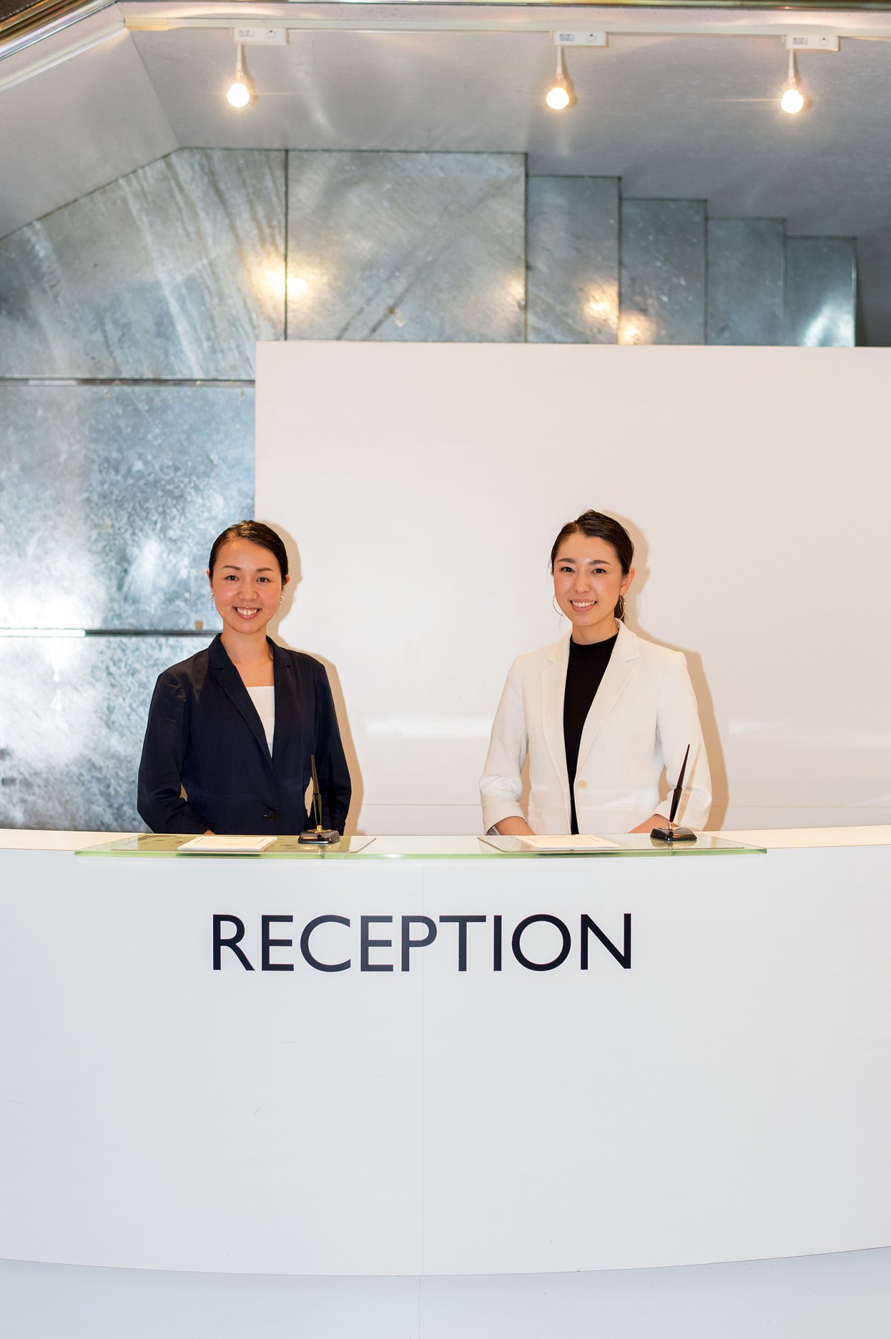 Two smiling women stand behind a modern white reception desk labeled 'RECEPTION' in a bright lobby with pendant lights.