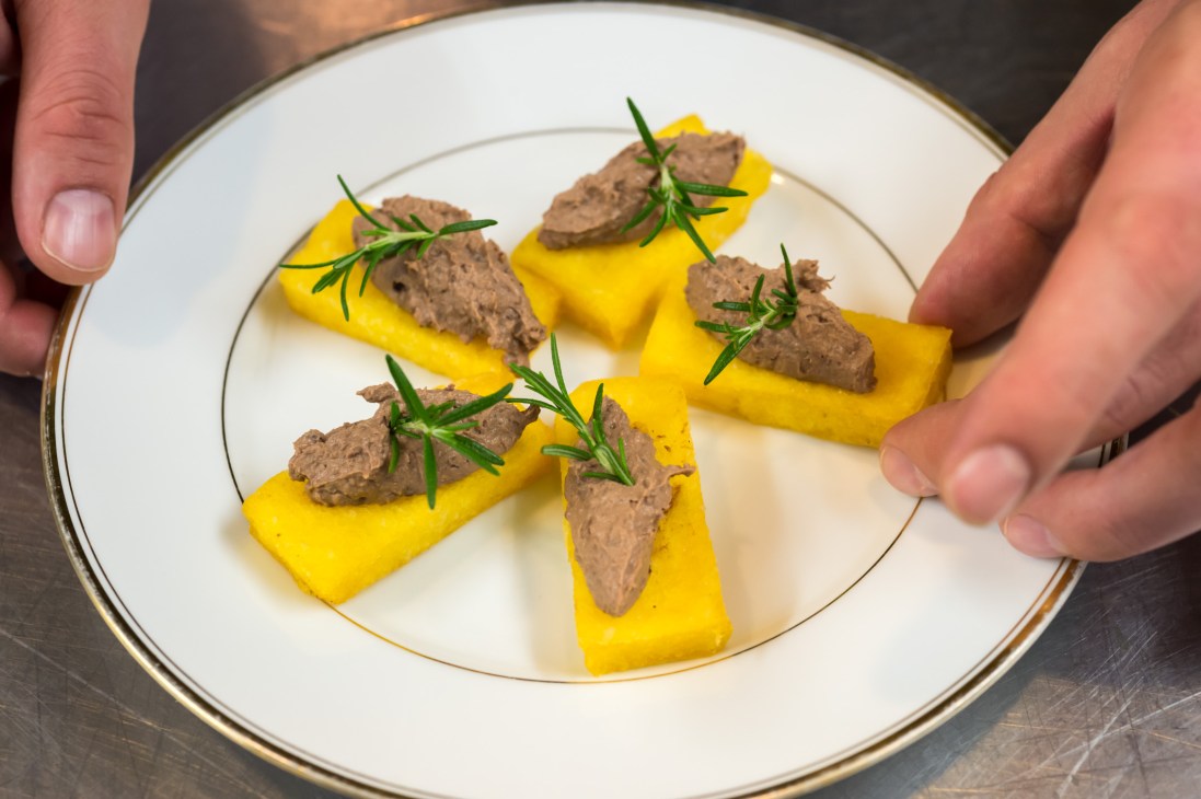 Hands presenting a white plate with fried polenta crostini topped with chicken-liver pâté and fresh rosemary garnish.