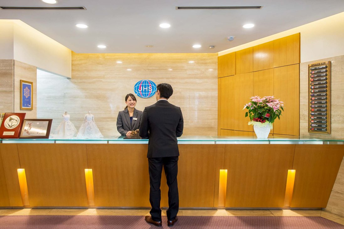 A receptionist serves a guest at the Japan Hotel School's training front desk with world clock visible.