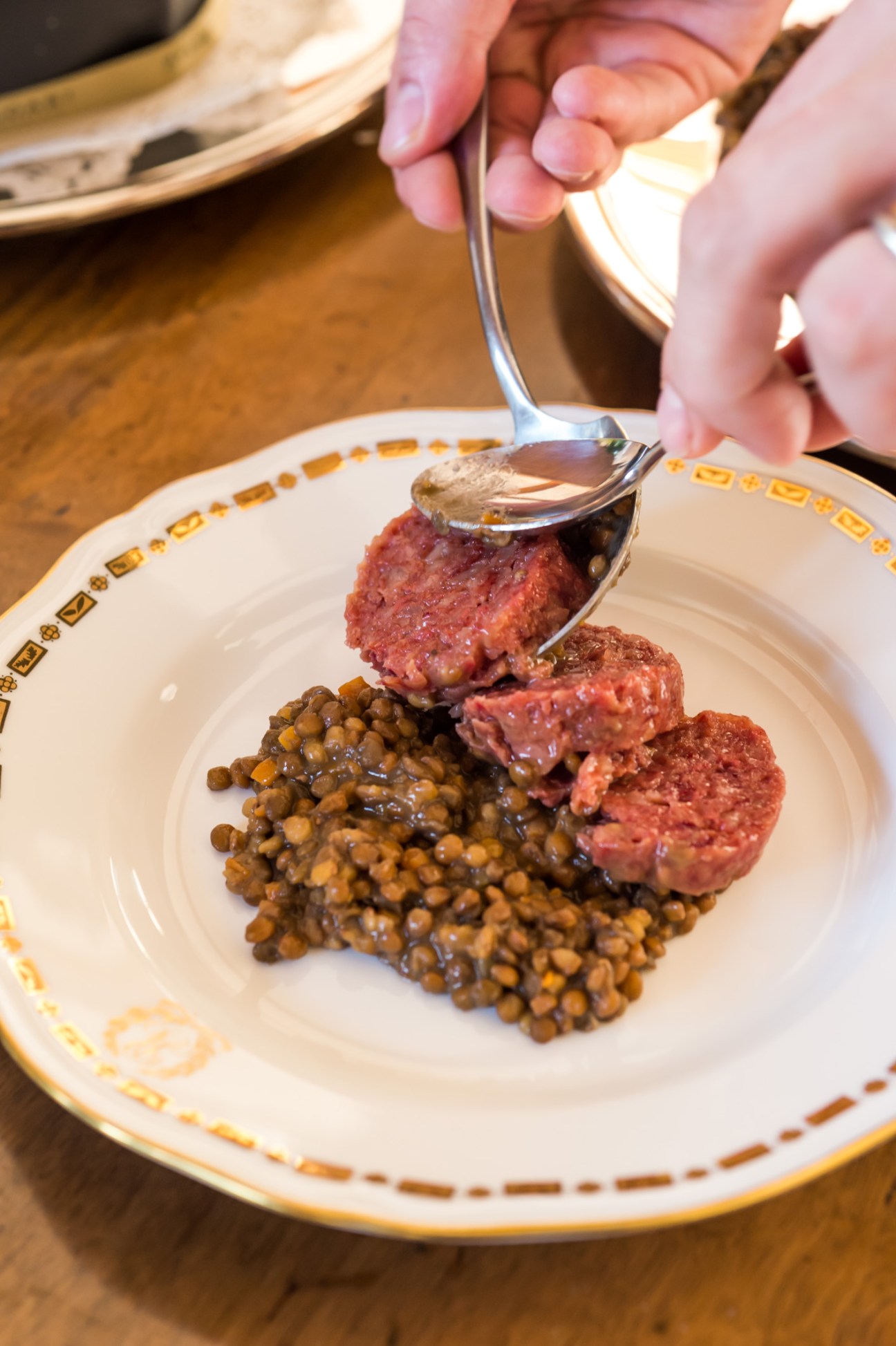 A hand holding a spoon serves zampone (stuffed pig's trotter) with lentils on a decorative plate.