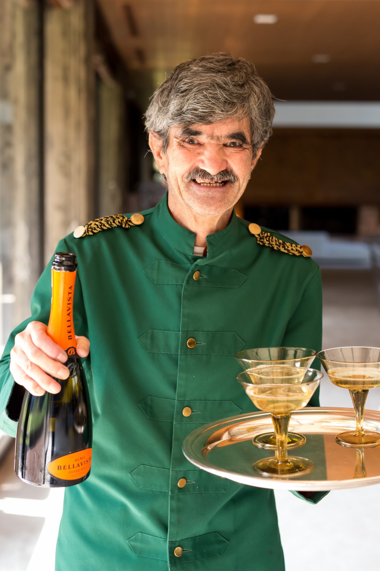 Man in green formal jacket holding Franciacorta sparkling wine bottle with glasses on a tray