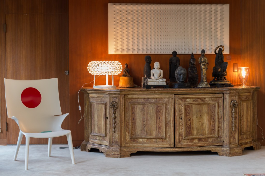 Buddhist statues in various materials displayed on a wooden cabinet against an orange wall with decorative lighting.
