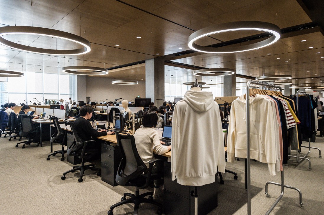 Open-plan office with employees at desks, clothing displayed on racks, and circular pendant lights overhead at Uniqlo City.