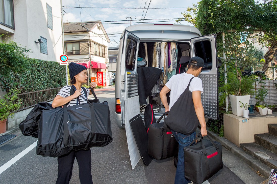 Two people loading garment bags and clothing into a white van during a Ginza magazine fashion shoot in a Tokyo residential street.