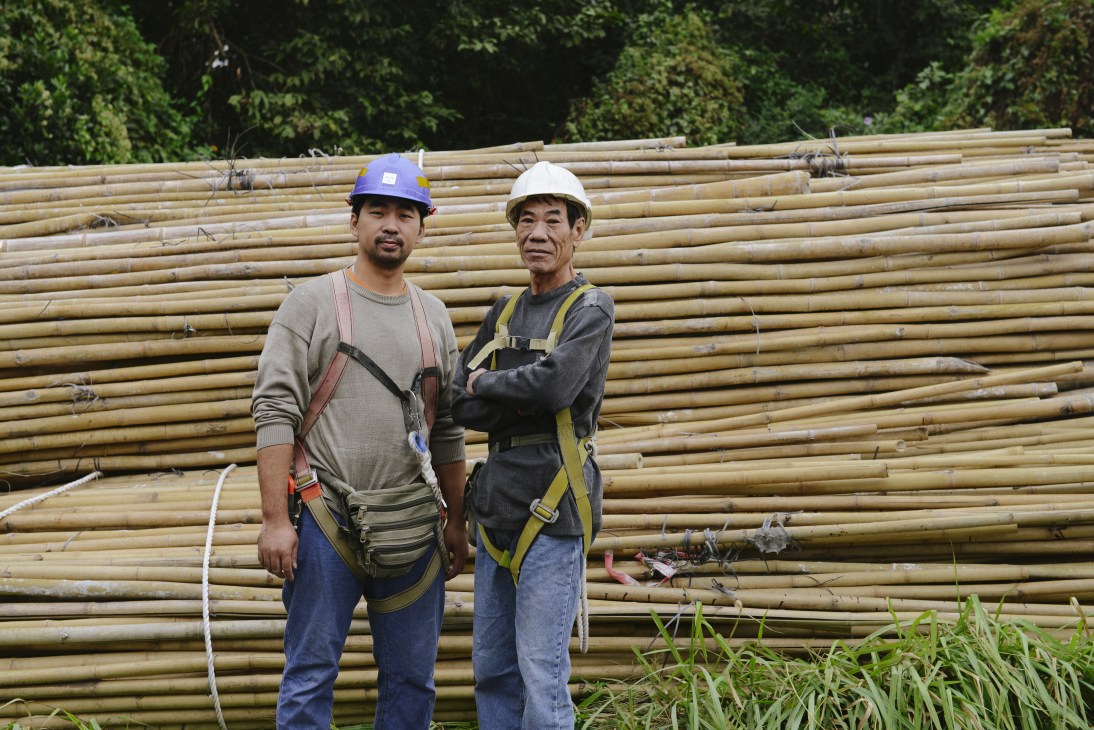 Two bamboo scaffolders Tang Woon Wing and Yu Wing Sung stand in front of stacked bamboo poles at a warehouse.